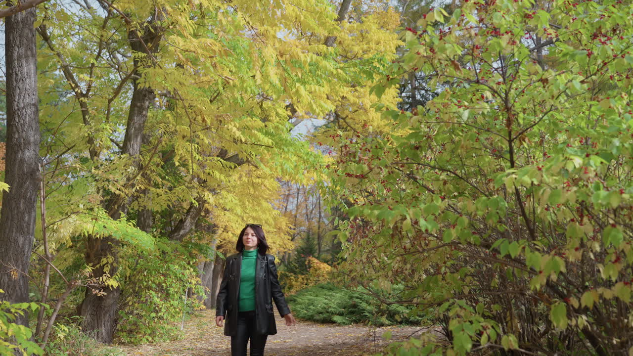 Mulher caucasiana caminhando por trilha de outono em floresta dourada, jaqueta de couro e suéter verde, passos lentos e firmes, sorriso leve, trilha frondosa, pouca profundidade, luz quente da tarde, humor contemplativo