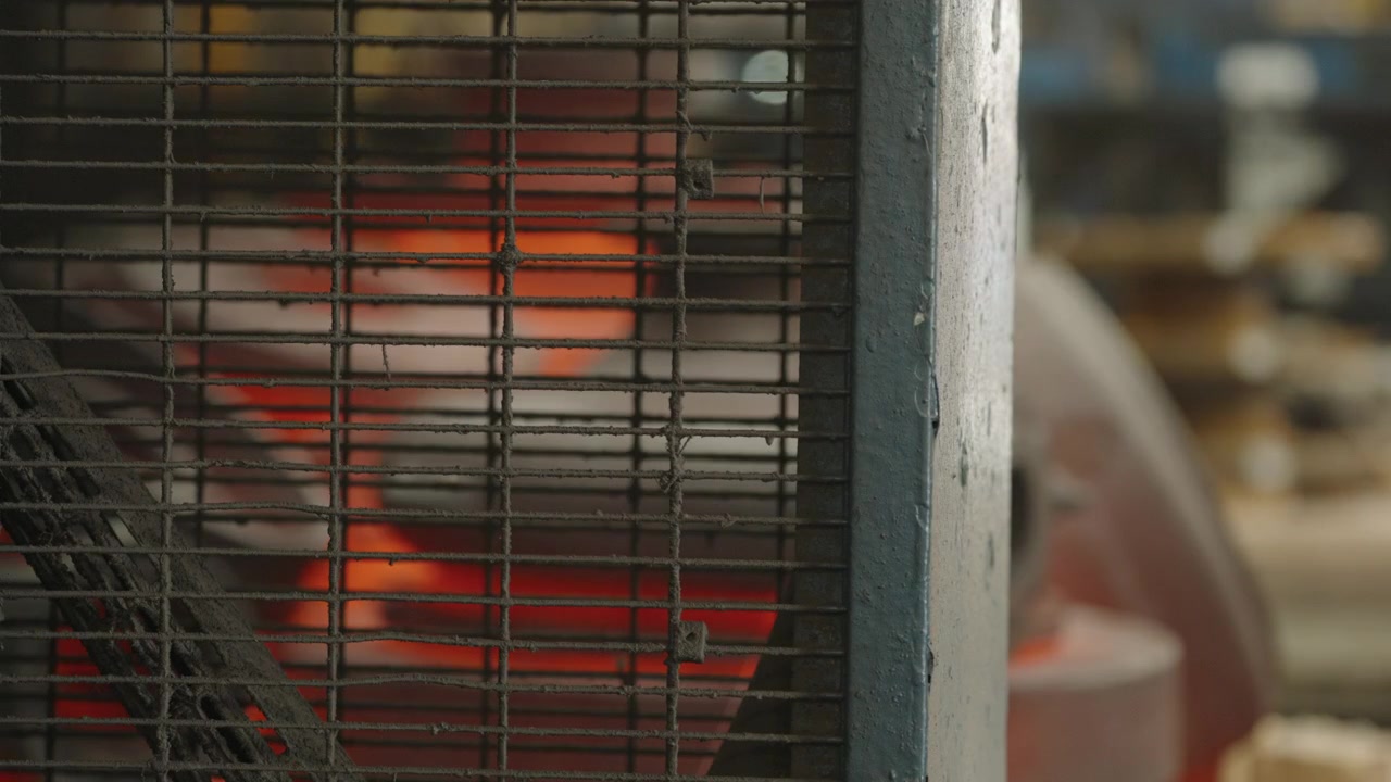 Metalventilatorgitter står foran en rødglødende ovn, med det varme industriværksted blødt sløret i baggrunden.