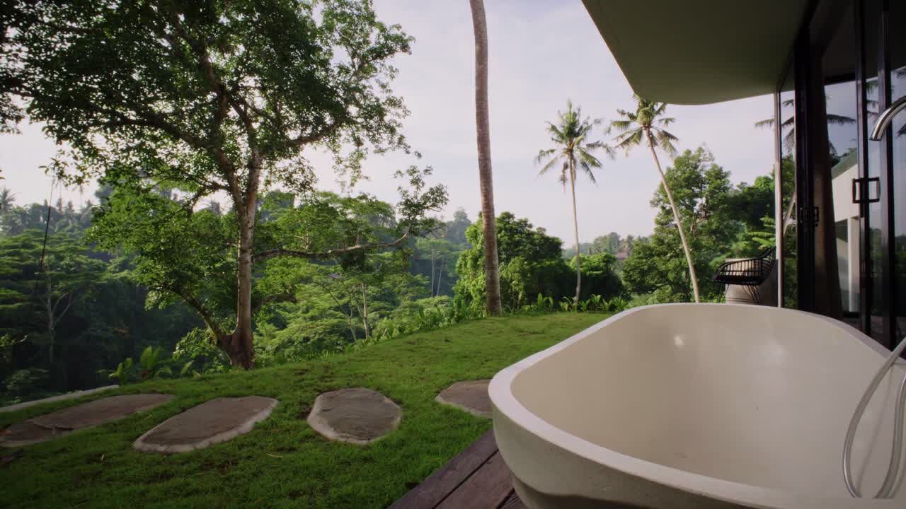 A white outdoor bathtub in a tropical backyard, surrounded by green plants and trees. It has a nice view of green hills and a coconut tree. a quiet and relaxing place to enjoy nature.