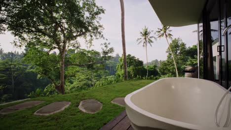 a white outdoor bathtub in a tropical backyard, surrounded by green plants and trees. it has a nice view of green hills and a coconut tree. a quiet and relaxing place to enjoy nature.