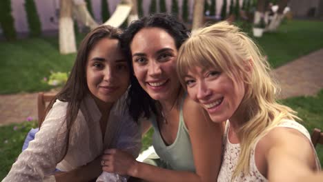 three joyful women met in veranda cafe outdoors taking selfie photo or video on smartphone, having fun, drink