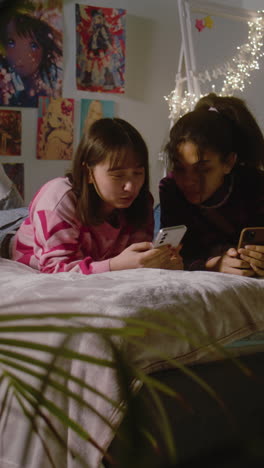 Vertical-shot-of-girl-sits-at-floor,-surfs-internet-using-tablet.-Mongolian-teen-lies-on-bed,-watches-content-with-friend.-Multi-ethnic-girls-together-spend-leisure-time-at-home.-Friends-relationship.