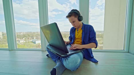 boy in headphones playing video game on laptop near the window. teenager gravely looking to the notebook screen pressing the keyboard. technology concept.