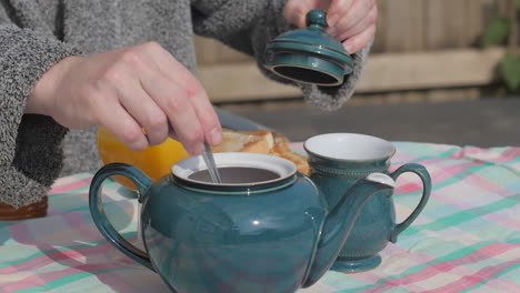 Die-Hand-Nimmt-Den-Deckel-Von-Der-Teekanne,-Um-Am-Frühstückstisch-Im-Freien-Umzurühren,-Zeitlupe