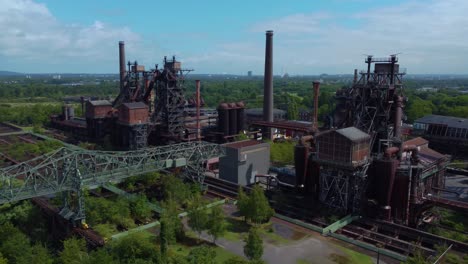 Rostige-Alte-Metallindustriefabrik-Mitten-Im-Wald-Mit-Mehreren-Abschnitten,-Schornsteinen,-Röhren-Und-Konstruktionen,-Die-Nebeneinander-Gebaut-Sind.-Vogelperspektive-Vom-Himmel,-Die-Sich-Von-Links-Nach-Rechts-Bewegt,-Sonnig