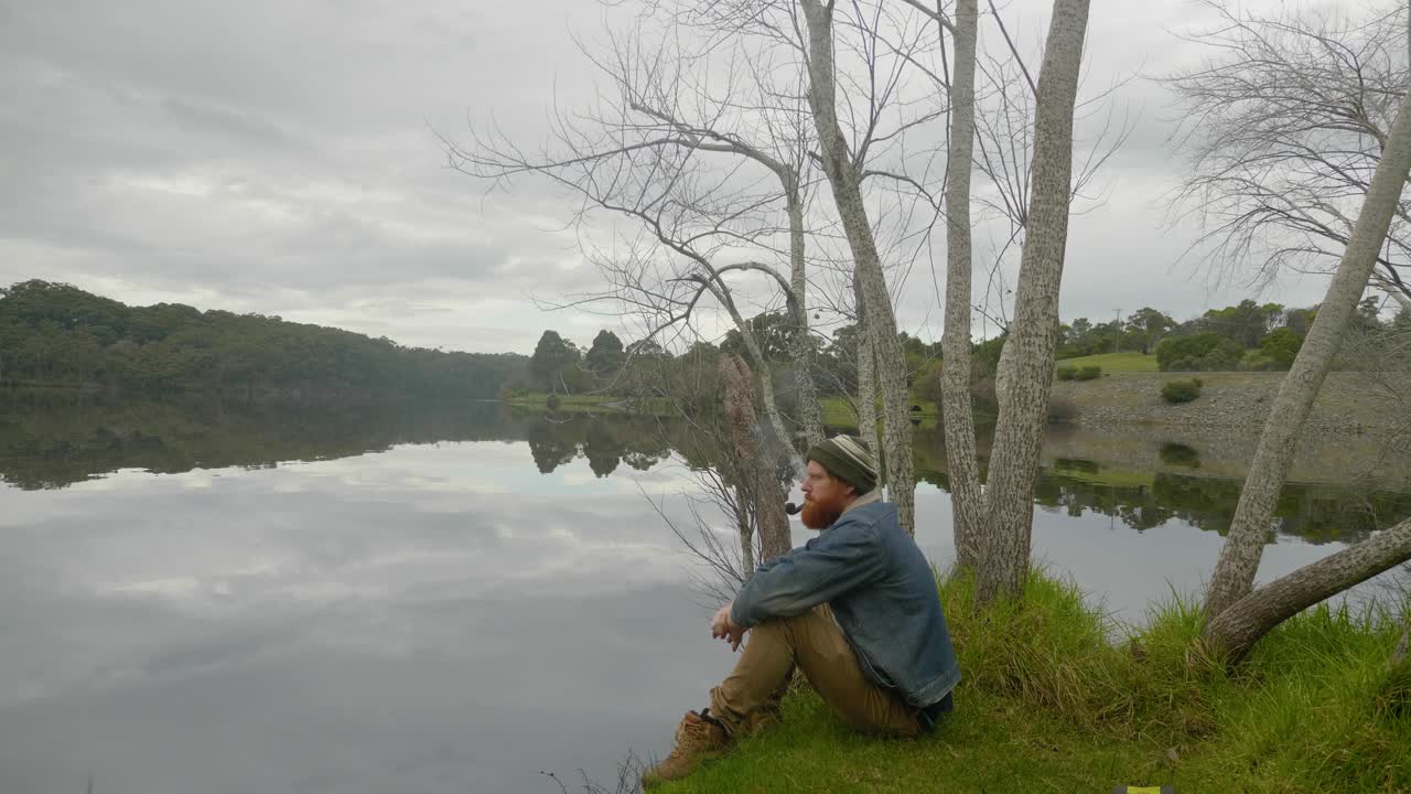una toma amplia de un pelirrojo barbudo sentado junto a un lago y fumando una pipa de tabaco
