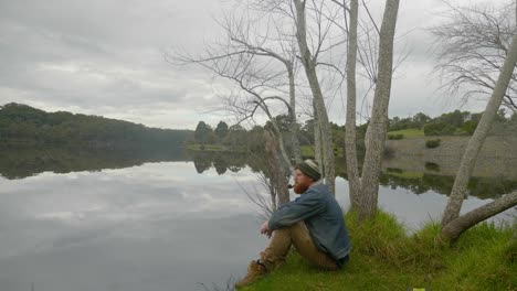 una toma amplia de un pelirrojo barbudo sentado junto a un lago y fumando una pipa de tabaco