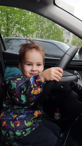 happy two-year-old toddler sitting on the driver's seat holding by the steering wheel. funny baby boy in a car. vertical video.