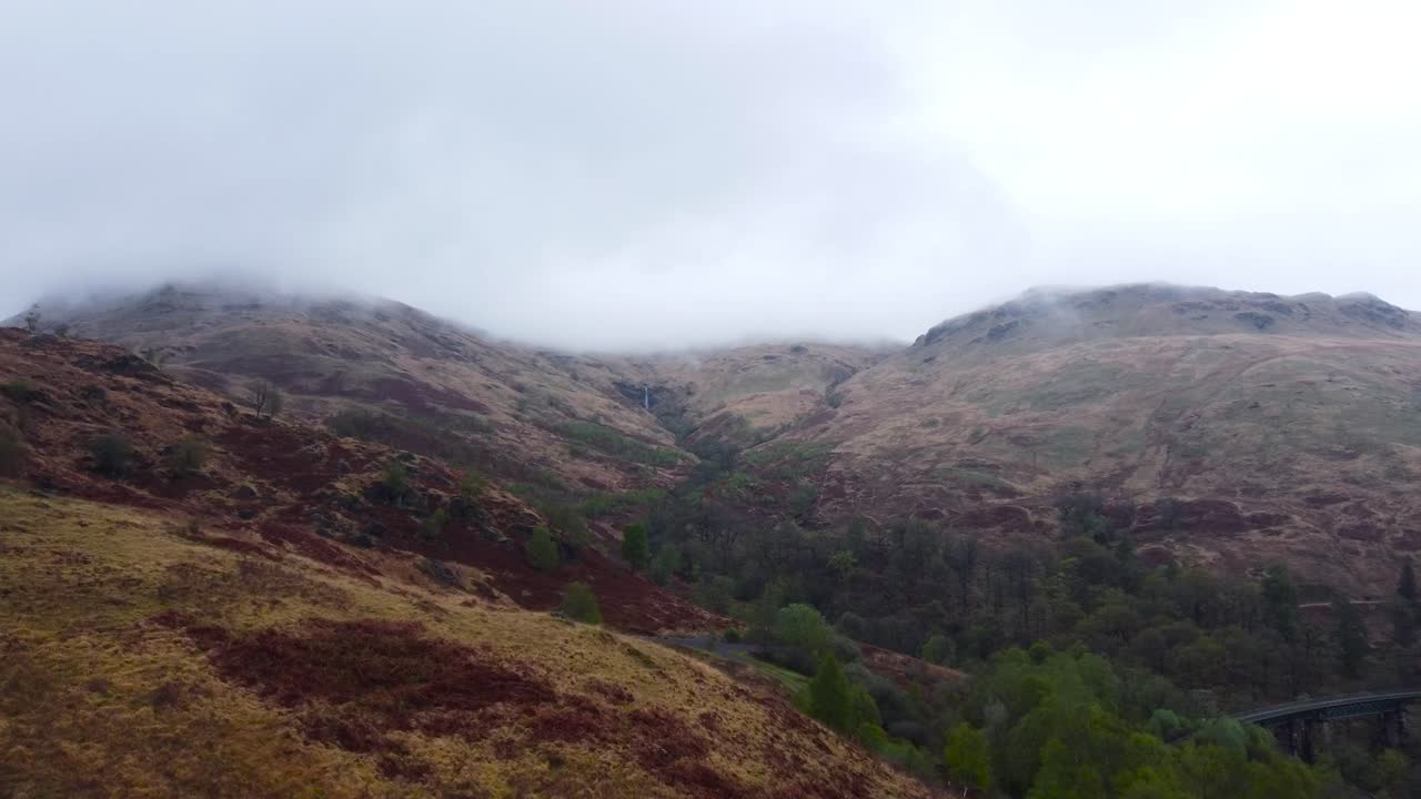 Aerial drone footage flying and gliding over misty brown and green nature covered Scotland terrain landscape that have hills and mountains with sloaps. Clouds are low to the mountains, between them.