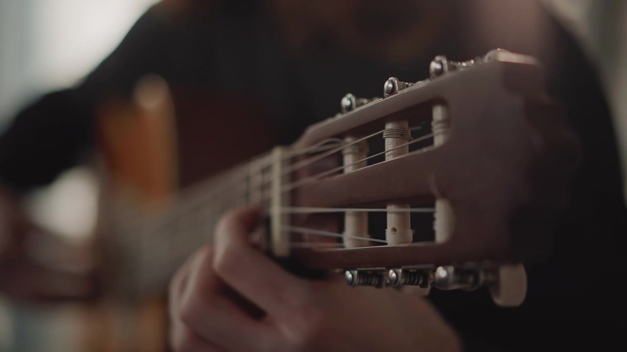 Primo piano della paletta di una chitarra classica, mani che premono le corde, un intimo istruttore di chitarra che dimostra una lenta tecnica fingerstyle in uno studio in penombra, retroilluminazione calda, profondità di campo ridotta, corde di nylon e esecuzione costante.
