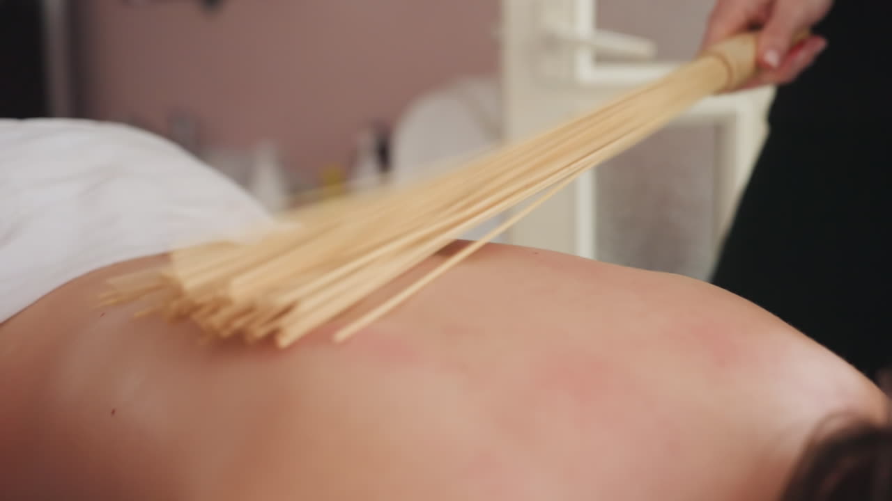 Close-up of fair skin woman receiving relaxing massage with bamboo broom, showing gentle strokes across her back while background remains softly blurred in warmly lit indoor spa setting