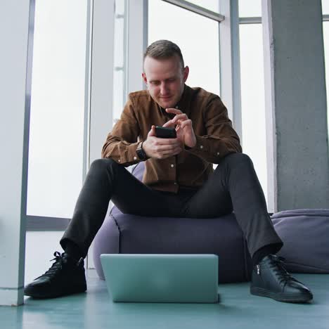 young man with gadgets on windows background. handsome guy is sitting on ottoman in office and using mobile phone. laptop on the floor. freelance concept