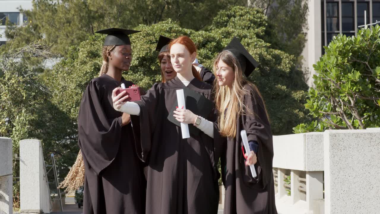 Impulsadas por su smartphone, diversas graduadas con togas caminando por una rampa del campus encuadrando una selfie grupal