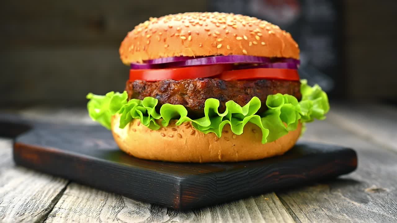 Hamburger appena fatto con lattuga, pomodoro e cipolla su un tagliere di legno. Un hamburger su un tagliere di legno con lattuga, pomodoro e cipolla impilati all'interno di un panino con semi di sesamo.