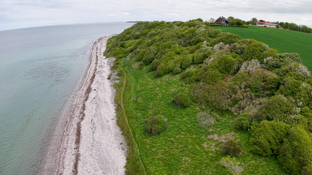Drone view of a rugged Danish coastline with white-pebble beach and lush green bluffs meeting the sea