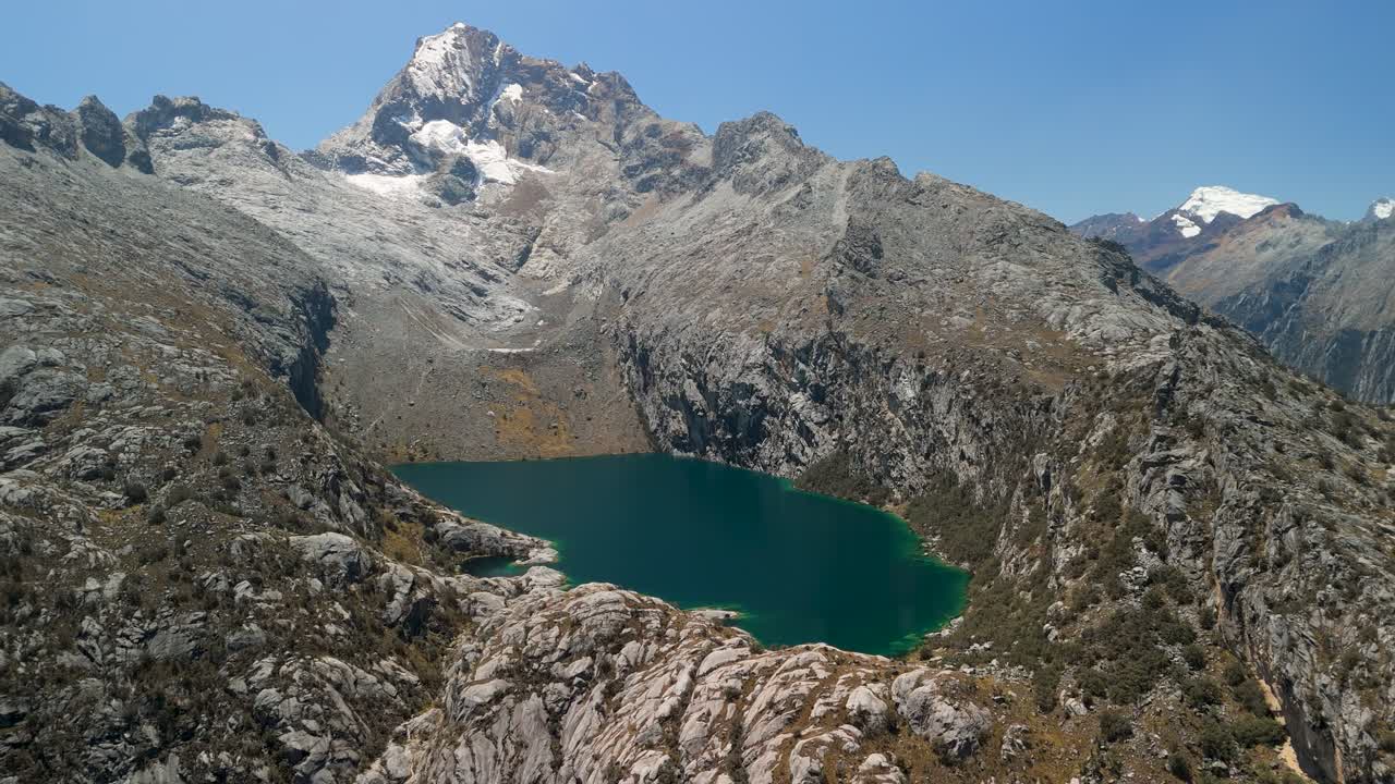 Drone pulls back to reveal stunning turquoise Laguna Churup surrounded by rugged Andes mountains under a clear blue sky in Peru