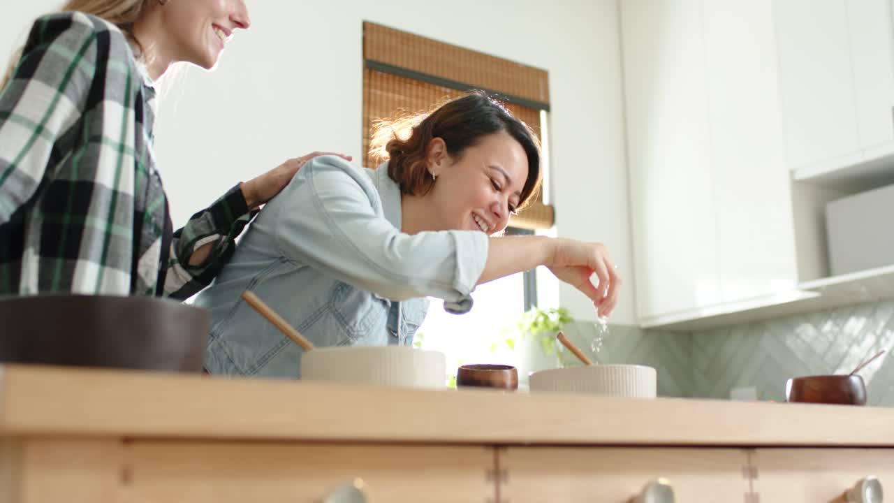 家のカウンターで食事の準備をする多様な女性の友人たち。女性は木のボウルから塩を振りかけている。