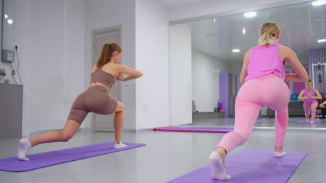 back view of two athletes in pink and brown workout gear stretching on yoga mats in a gym, mirror reflection captures synchronized movements as they perform balance and flexibility exercises