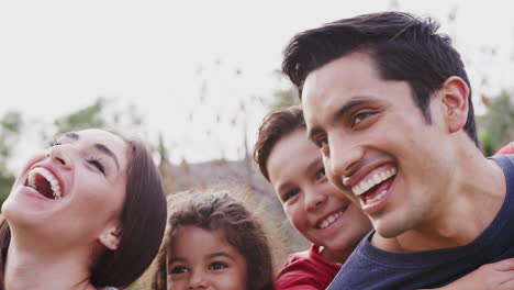 Head-and-shoulders-close-up-of-smiling-young-Hispanic-parents-piggybacking-their-children-in-park