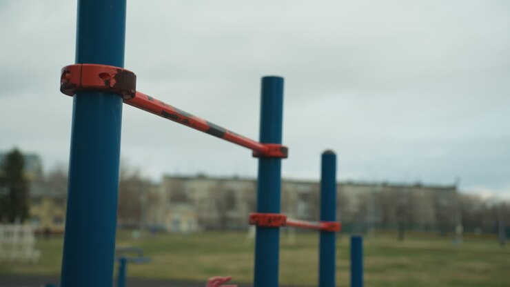 Two boys are working out on pull-up bars in an outdoor gym area, each gripping the bars but appearing tired and struggling to continue their exercises
