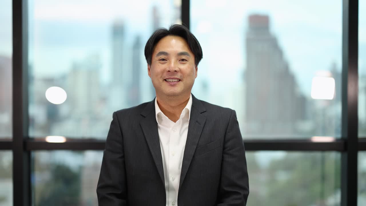 Confident Asian businessman bows with wai greeting, smiling in bright office with cityscape background