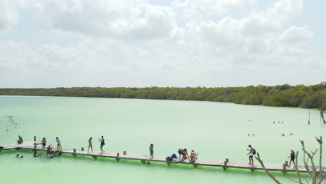 Fly-over-people-relaxing-at-water,-sitting-at-long-wooden-pier-or-bathing-in-natural-swimming-pool.-Kaan-Luum-lagoon,-Tulum,-Yucatan,-Mexico