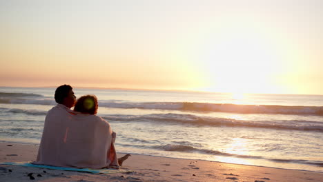 Pareja-Birracial-Envuelta-En-Una-Manta,-Viendo-Una-Puesta-De-Sol-En-La-Playa-Con-Espacio-Para-Copiar