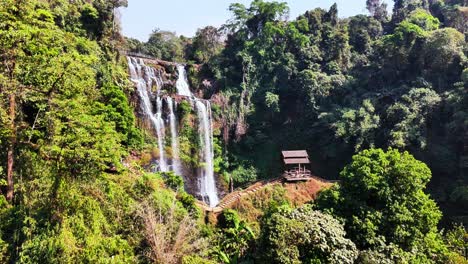 static camera shot of tad gneuang waterfall plunging through verdant jungle in champasak province, laos, with a wooden pavilion beside the falls offering a serene viewpoint amid lush greenery