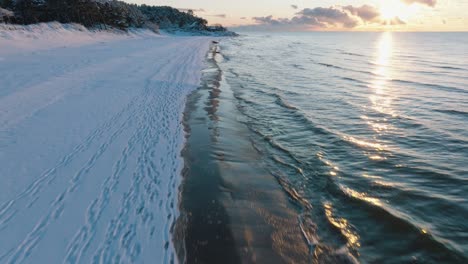 Aerial-footage-of-beach-covered-with-snow,-sunny-winter-day-at-the-sunset,-golden-hour,-Nordic-woodland-pine-tree-forest,-Baltic-sea-coast,-wide-drone-shot-moving-forward-low