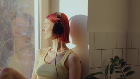 relaxed girl in headphones listening to music on windowsill at home