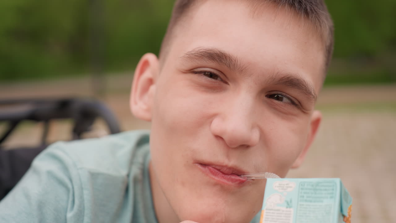 Caucasian Youth Enjoying Summer Drink, Male Individual With Playful Wink And Juice In Park Setting, Young Caucasian Male Smiling And Sipping Juice While Seated On Park Bench During Summer