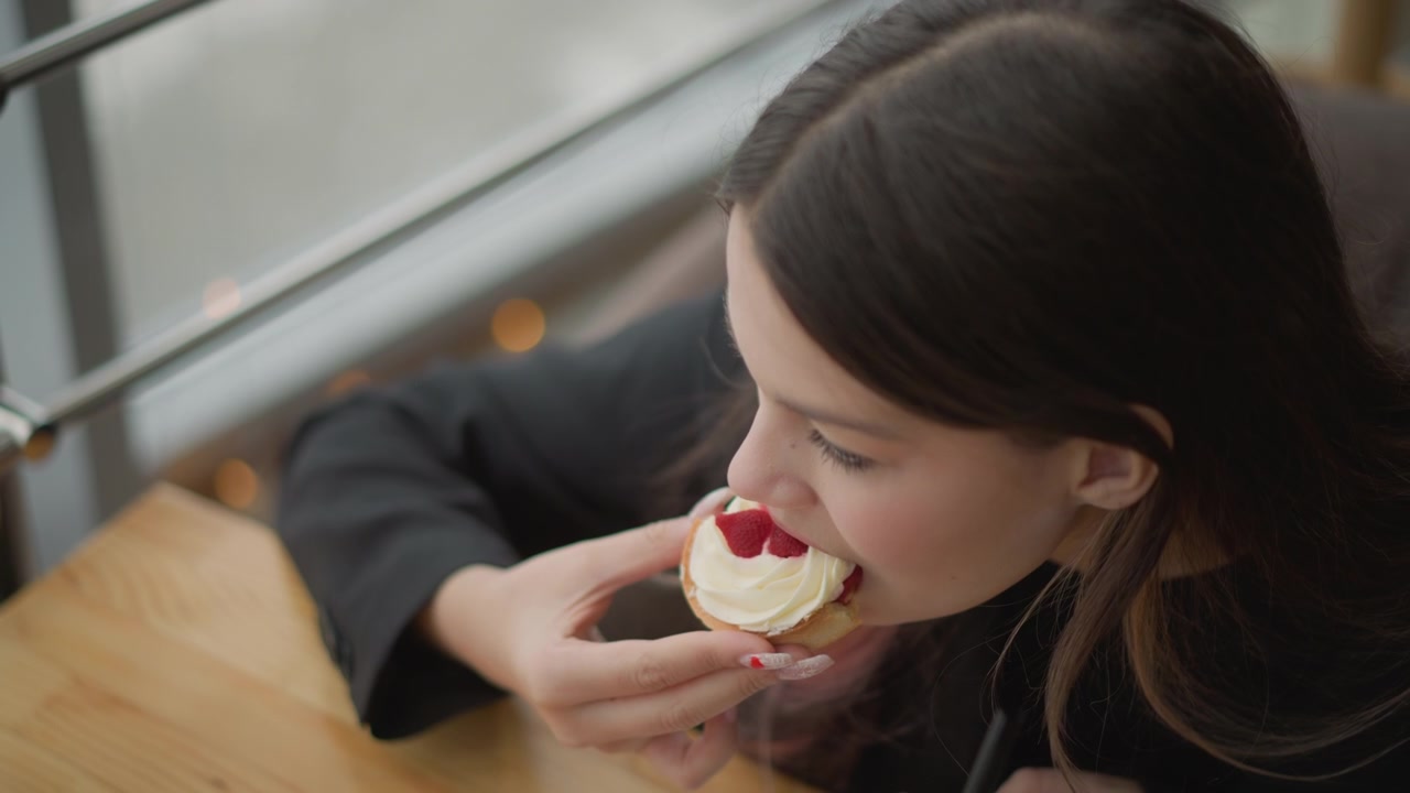 Young woman leans over wooden table beside window and brings small heart topped pastry to her mouth, biting flaky crust with both hands in soft natural light for relaxed indoor snacking