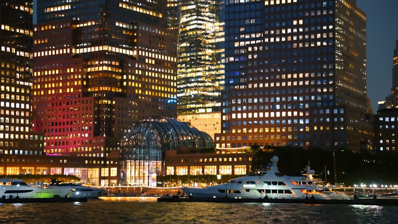 Luxury yachts docked near illuminated skyscrapers in New York. Bright lights of New York skyscrapers reflect on the water where luxury yachts are docked