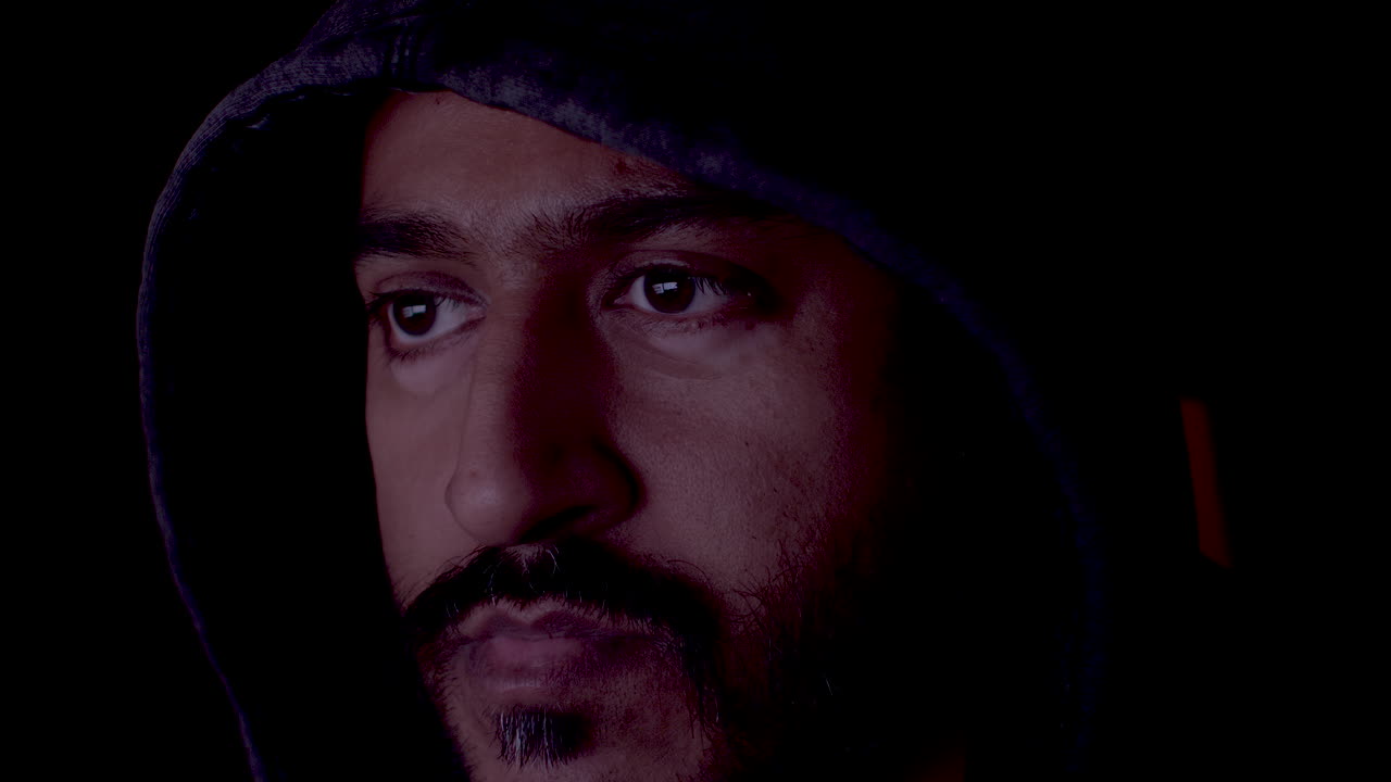 A face of a brown-eyed man wearing a hoodie. Man is exploring the screen and browsing the internet in front of his desktop. The screen light reflects on the man's face