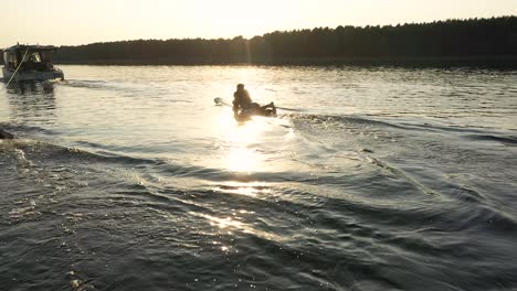 Surfer-lies-on-surfboard-behind-boat-with-sunset