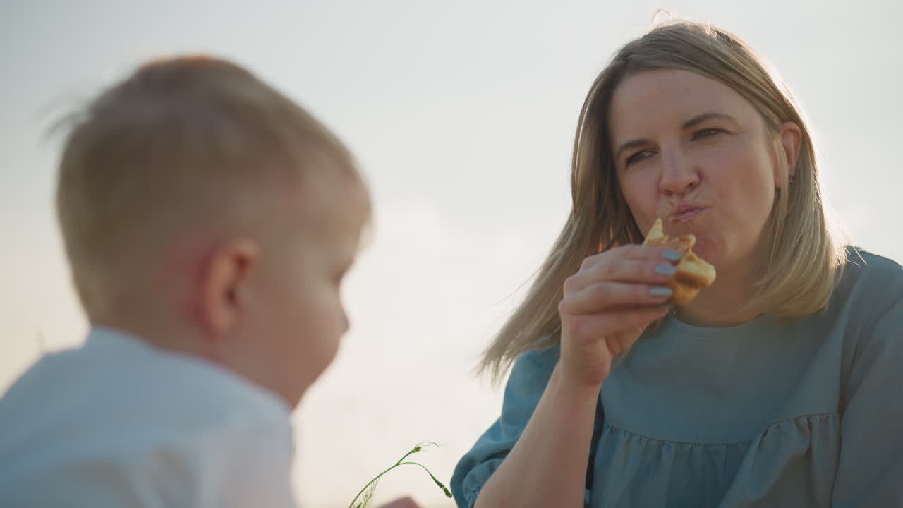 A little boy in a white shirt sits in a grassy field, sharing a hotdog with his mother, who is dressed in blue. She takes a bite and hands it back to him, capturing a tender moment of sharing