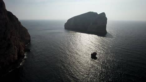 Toma-Aérea-De-La-Silueta-De-La-Enorme-Formación-Rocosa-Contra-El-Sol-Que-Se-Refleja-En-La-Superficie-Del-Mar