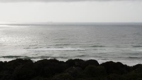 Eine-Lange-Einstellung-Von-Wellen,-Die-Auf-Einen-Dunklen-Strand-Schlagen,-Mit-Einem-Schiff,-Das-In-Der-Ferne-Kaum-Sichtbar-Ist