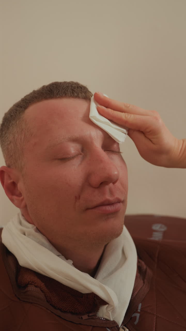 Uomo che riceve una terapia facciale con microcorrenti, l'estetista deterge delicatamente la fronte e applica cuscinetti conduttivi, asciugamano drappeggiato attorno al collo, primo piano di un'espressione rilassata, movimenti di massaggio delicati.