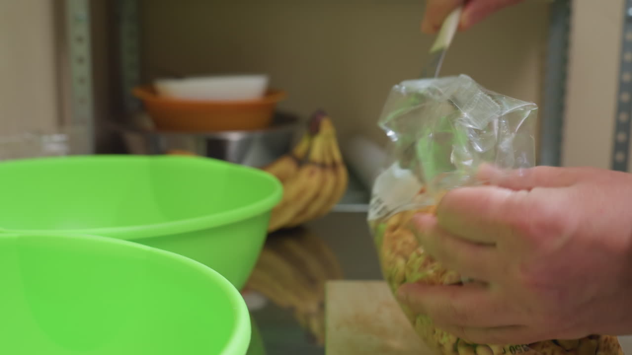 two bright green bowls on countertop, pantry assistant opening bag of granola and preparing portions for breakfast with bananas in background, measured pouring motion and casual kitchen organization
