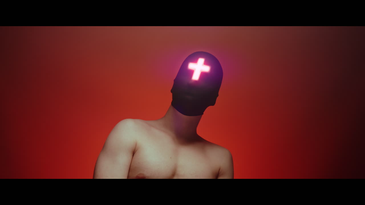 Masked and hooded man with cross of light glowing on his face looks in the camera - red background