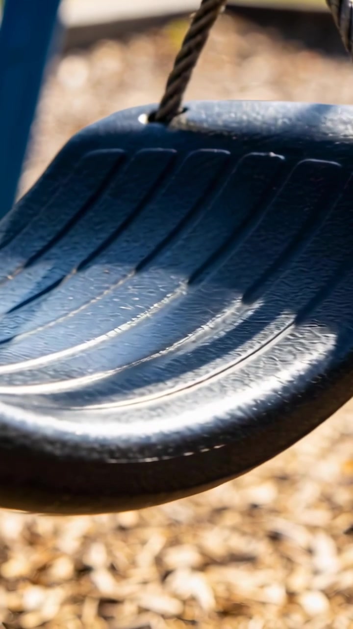 Video verticale: Brezza che muove delicatamente un sedile di altalena nero, che si inclina nel parco giochi, corda intrecciata, creste illuminate dal sole