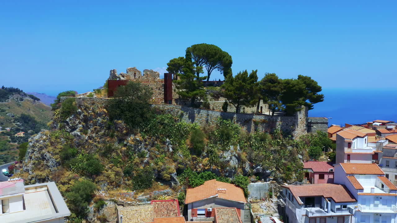 Drone flying around the remains of an old castle on the small rocky cliff surrounded by the Sicilian village; sea in the background. Castelmola, Italy