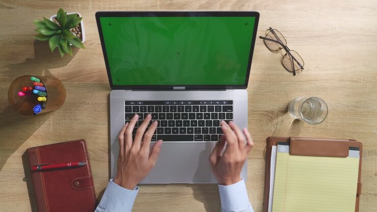 Draufsicht auf einen Laptop-Computer mit Mockup-Greenscreen-Display auf einem Holzschreibtisch, daneben ein Notizbuch mit Stiften, Brille und einem Glas Wasser. Langsames Herauszoomen, Nahaufnahme.