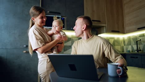 happy caucasian man sits in front of laptop. man turns to his kid in mom's hands rejoicing about some news on the screen.