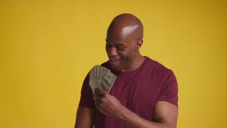 studio shot of excited mature man celebrating winning cash prize holding handful of 100 dollar bills against yellow background