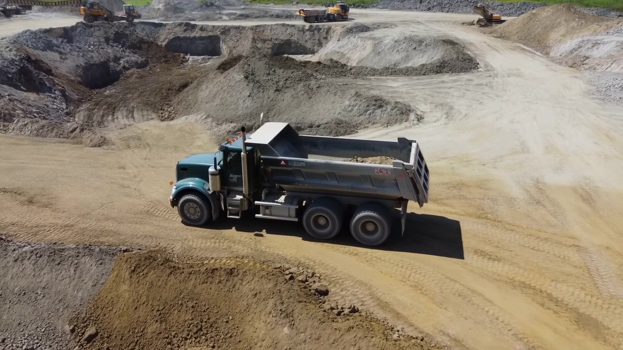 Oblique aerial tracking view follows loaded dump truck moving through broad excavation site as other heavy machines, layered soil, gravel piles, cut slopes, and tire-marked roads fill background.