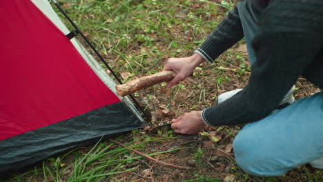 aerial view shows man squatting near red tent securing corner by hammering peg into forest ground using thick wooden stick surrounded by pine needles grass and scattered leaves during camping setup