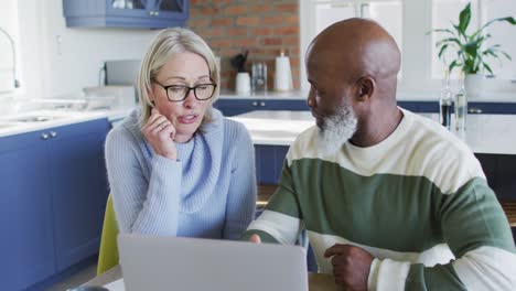 una pareja diversa estresada en la cocina sentada en la mesa, usando una computadora portátil