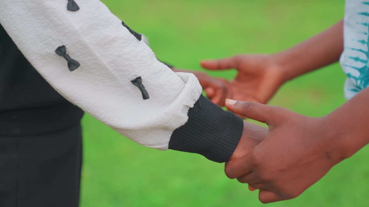 Black mother guiding child handhold across grass, supportive grip as child takes step, outdoor teaching scene with gentle encouragement, casual sweater sleeve and vivid green lawn background